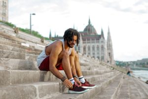 Selective Focus Photo of Man in White Vest and Red Shorts Tying His Shoes While Sitting on Concrete Stairs
